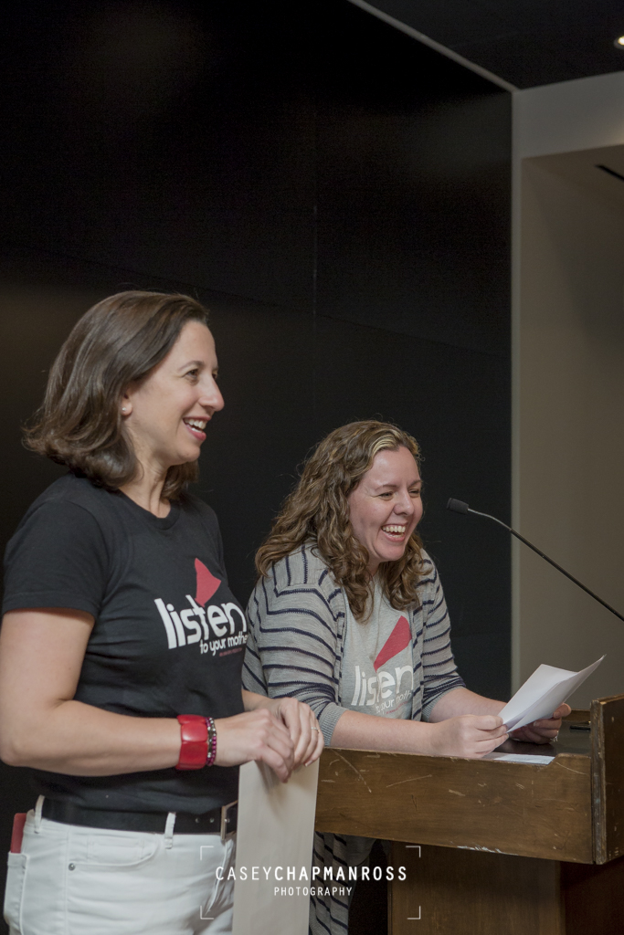Two women in Listen To Your Mother T-shirts at podium laughing